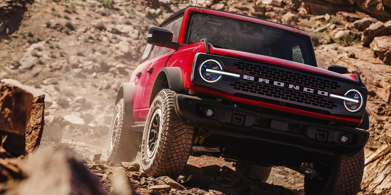 The image features a red Ford Bronco tackling rough, rocky terrain, showcasing its rugged, off-road capabilities and bold design in a challenging natural environment.